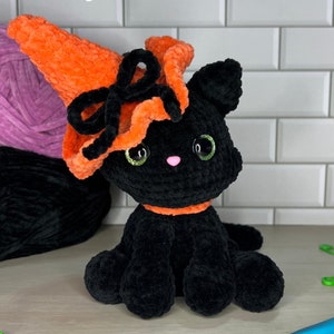 Può includere: Un gatto di uncinetto nero che indossa un cappello da strega arancione con un fiocco nero. Il gatto è seduto su una superficie di legno con gomitoli di lana sullo sfondo. Il testo "Digital Pattern" è in alto nell'immagine e "PATTERN BY @MYCRAFTYAUNTIE" è in basso.