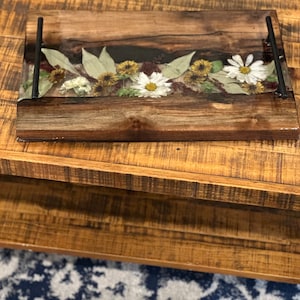 May include: A rectangular wooden serving tray featuring a central clear resin inlay embedded with pressed white daisies, yellow wildflowers, and green leaves. The tray has a dark wood grain finish and two matte black metal handles on the sides.