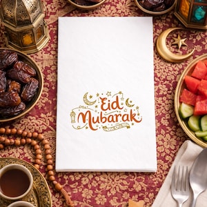May include: A white paper napkin with the words "Eid Mubarak" in orange script, decorated with crescent moons, stars, and a lantern. The napkin is surrounded by dates, fruit, and traditional decorations on a patterned red tablecloth.