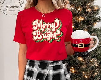 May include: Red t-shirt with a green and white "Merry & Bright" graphic on a leopard print background. The shirt is being worn by a person with a black and white plaid skirt.