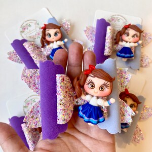 May include: Purple and white glitter hair bow with a blue and white dress doll charm. The doll has brown hair, brown eyes, and a red bow.