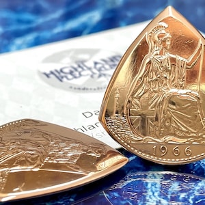 Puede incluir: Dos monedas triangulares doradas con un relieve detallado de una figura sentada, posiblemente Britannia, y el año 1966. Las monedas están sobre una superficie azul y blanca, con una tarjeta parcialmente visible en el fondo.