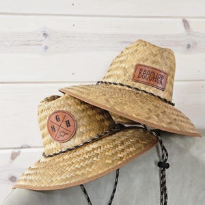 May include: Two straw hats with brown leather patches. One hat has the word "BEECHER" on a rectangular patch. The other hat has a circular patch with the letters "G", "M", and "H" and a small graphic. Both hats have a black and white rope around the brim.