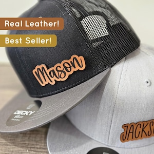 Peut inclure: Deux casquettes de baseball noires et grises avec des patchs en cuir. La casquette du haut a un dos en maille noire et un patch en cuir brun avec le nom "Mason" en cursive. La casquette du bas a un devant gris et un patch en cuir brun avec le nom "JACKS" en cursive.