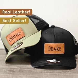 May include: Two baseball caps with leather patches. The first cap is green with a black mesh back and a brown leather patch that says "Oliver". The second cap is black with a brown leather patch that says "Drake".