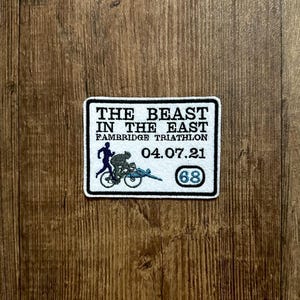May include: White patch with blue and black embroidery. It reads "The Beast in the East" with the text "Cambridge Triathlon" and "04.07.21" below. A number "68" is in a blue circle.