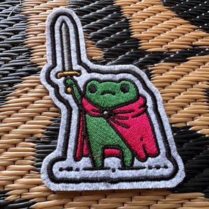 May include: An embroidered patch featuring a green frog wearing a bright pink cape and holding a silver sword with a gold hilt. The patch has a white border and a dark outline, showcasing detailed stitching throughout the character design.