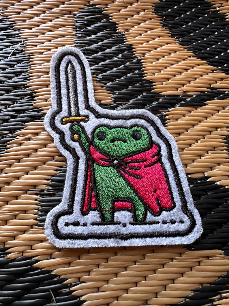 May include: An embroidered patch featuring a green frog wearing a bright pink cape and holding a silver sword with a gold hilt. The patch has a white border and a dark outline, showcasing detailed stitching throughout the character design.