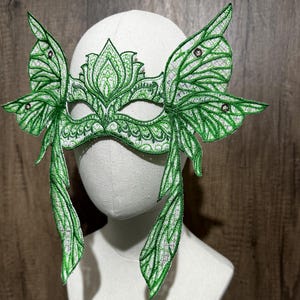 May include: Green masquerade mask with butterfly wings and intricate floral details. The mask is made of lace and has a delicate, feminine design.