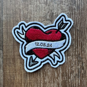 May include: A blue embroidered patch featuring a red heart pierced by an arrow and a white banner with the text "12.05.24".