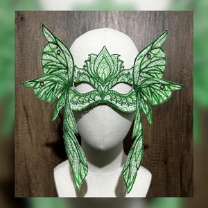 May include: A green masquerade mask with butterfly wings and intricate details. The mask is made of fabric and has a white base. It is decorated with green and silver embroidery and rhinestones.
