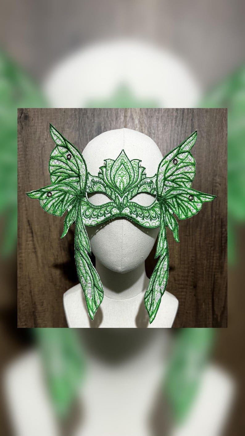 May include: A green masquerade mask with butterfly wings and intricate details. The mask is made of fabric and has a white base. It is decorated with green and silver embroidery and rhinestones.