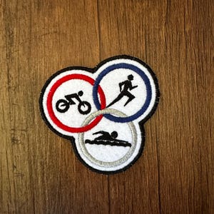 May include: A white, red, blue, and silver embroidered patch with three interlocking circles. The circles depict a swimmer, a cyclist, and a runner.