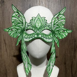 May include: A green masquerade mask with butterfly wings and intricate floral designs. The mask is made of fabric and has a white background.
