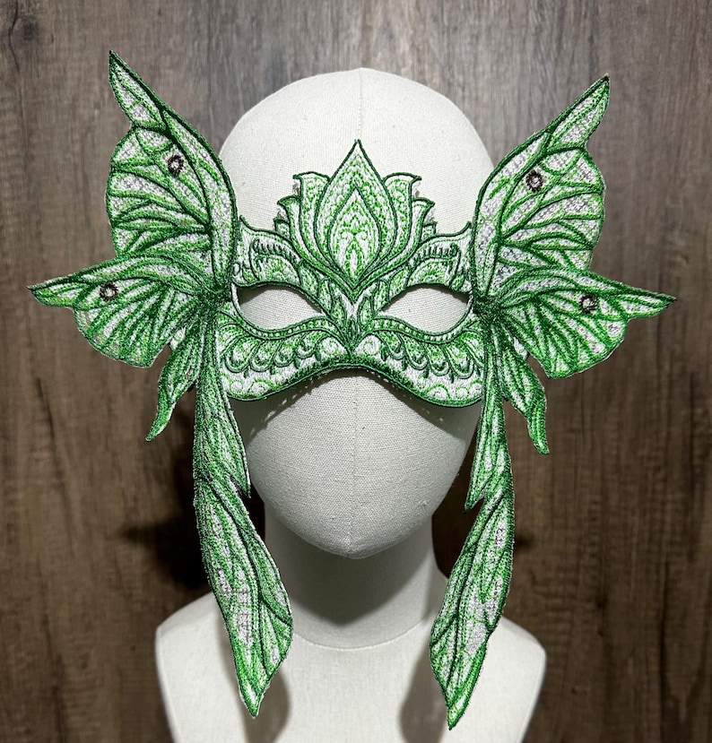 May include: A green masquerade mask with butterfly wings and intricate floral designs. The mask is made of fabric and has a white background.
