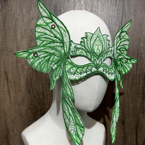 May include: A green masquerade mask with butterfly wings. The mask is made of lace and has a delicate floral design. The wings are detailed with intricate patterns and have a shimmery finish.