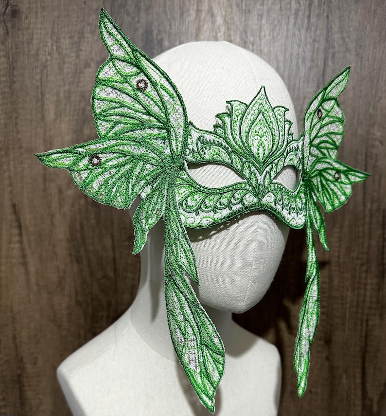 May include: A green masquerade mask with butterfly wings. The mask is made of lace and has a delicate floral design. The wings are detailed with intricate patterns and have a shimmery finish.