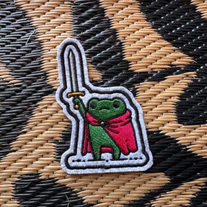 May include: An embroidered patch featuring a green frog wearing a bright pink cape and holding a silver sword aloft. The design has a thick white border and black outlines, depicting the frog in a standing position against a light gray background.