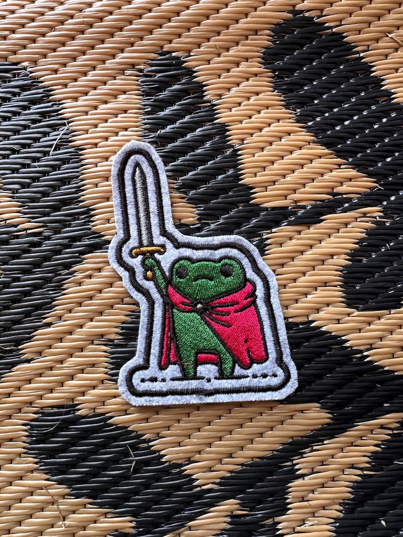 May include: An embroidered patch featuring a green frog wearing a bright pink cape and holding a silver sword aloft. The design has a thick white border and black outlines, depicting the frog in a standing position against a light gray background.
