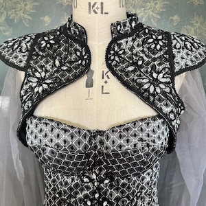 Könnte beinhalten: Ein schwarz-weiß gemustertes Bustierkleid mit passendem Bolero. Der Bolero hat eine Blumenstickerei und einen hohen Kragen. Das Kleid wird auf einer Schaufensterpuppe präsentiert und zeigt das komplizierte Design und die Schneiderkunst des Kleidungsstücks.
