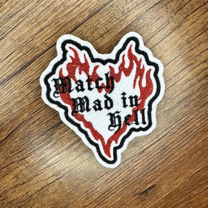 May include: A white patch with a black border and the words "Match Mad in Hell" in gothic font. Red flames surround the text, creating a heart-like shape. The patch is on a wooden surface.