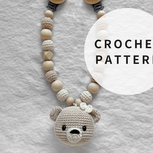May include: A crocheted bear head with a white flower attached to the top, hanging from a wooden and crocheted bead chain with a clip on one end. The chain is on a white surface. The text "CROCHET PATTERN" is in a white circle on the image.