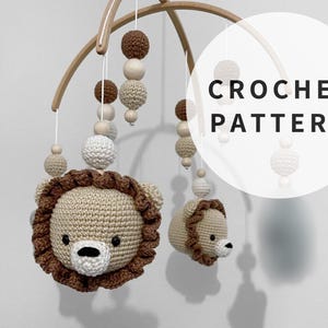 Op de afbeelding: Een houten babymobiel met gehaakte leeuwenkoppen, bruine en witte ballen en houten kralen. De mobiel hangt aan een wit plafond. De tekst "CROCHET PATTERN" staat in zwart op een witte cirkel.