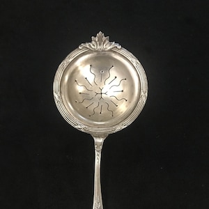 Peut inclure: Une passoire à thé en argent avec un bol rond perforé et un long manche. Le bol a un bord décoratif et un motif de lignes courbes rayonnant du centre. La poignée présente des motifs floraux et de volutes.