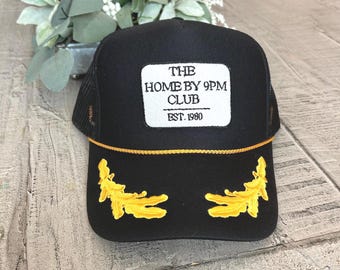 May include: Black trucker hat with a mesh back and a white rectangular patch. The patch reads "THE HOME BY 9PM CLUB EST. 1980" in black text. The hat features gold embroidered leaf designs and a gold rope detail.