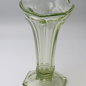 May include: A pale green glass vase with a fluted body and a hexagonal base. The vase has a scalloped rim and a slender, elegant shape. The glass has a slight tint, giving it a vintage appearance.