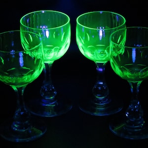 May include: Four green-tinted glass goblets with a decorative pattern on the bowl and stem. The glasses are illuminated, creating a glowing effect against a dark background. The goblets are likely for serving beverages.