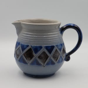 Può includere: Una brocca in ceramica con corpo grigio chiaro e texture a coste orizzontali. Il centro è decorato con una fascia di rombi blu scuro e grigio antracite. Presenta un manico curvo e un beccuccio per versare con una finitura liscia.