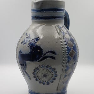 Op de afbeelding: Een grijze stenen kan met een blauw geglazuurd springend hert en een bloemmotief in medaillonstijl. Het zijpaneel toont een blauw en grijs ruitpatroon, met blauwe horizontale banden rondom de hals en de voet van de kan.