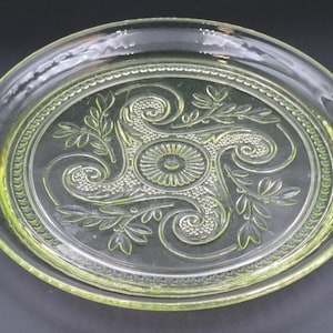 May include: A clear glass serving dish with a decorative floral and swirl design. The dish has a round shape with a slightly raised rim and a central oval area with embossed detailing. The glass has a slight green tint.