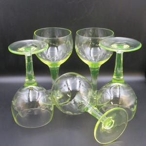 May include: Five clear glass wine goblets with a bright green stem and base. The glasses have a rounded bowl and a decorative etched floral pattern. The goblets are arranged on a dark background.