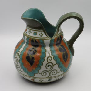 May include: A ceramic jug with a white base, featuring a repeating pattern of orange scalloped shapes, teal borders, and dark blue leaf motifs. The handle and interior rim are a solid olive green, and the exterior includes small dot accents.
