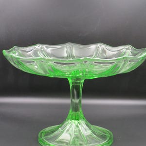 May include: A clear, green glass pedestal serving dish. The dish has a scalloped rim and a round base. The stem is fluted and the glass has a slight glow. The dish is likely vintage and suitable for serving desserts or fruit.