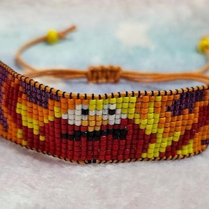 May include: A colorful beaded bracelet with a cartoon face design. The bracelet is made with orange, yellow, red, purple, and white beads. The bracelet is tied with a brown cord.
