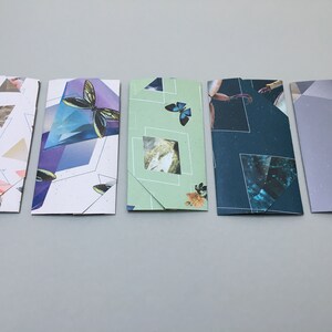 May include: Five folded cards with different designs. The cards are all white with different patterns and colors. The first card has a geometric pattern with triangles in shades of pink, gray, and white. The second card has a geometric pattern with blue, purple, and white triangles. The third card has a green background with a butterfly and a square window with a picture of a rock formation. The fourth card has a dark blue background with a crab and a geometric pattern with blue, white, and gray triangles. The fifth card has a light purple background with a geometric pattern with gray triangles.