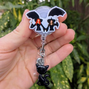 May include: A retractable badge reel featuring a witch-themed design. The top has a white background with two witch figures holding umbrellas. A black cat charm hangs below. The reel is attached to a silver-colored clip.