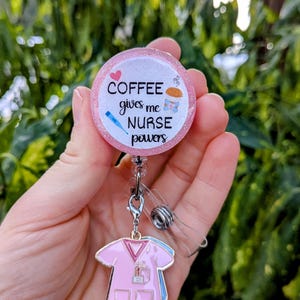 May include: Pink glitter badge reel with a pink and white circle that says "Coffee gives me nurse powers" with a pink heart and a coffee cup graphic. The reel has a pink and blue scrub top charm.
