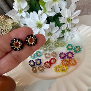 Puede incluir: Una colección de pequeños pendientes de botón circulares con cuentas facetadas dispuestas en un patrón floral alrededor de un anillo central de alambre dorado. Los pendientes están disponibles en varios colores, incluyendo negro, azul, rojo, morado, naranja, amarillo, verde y blanco.