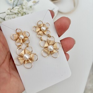 Puede incluir: Un par de pendientes de alambre dorado con detalles de perlas. Cada pendiente presenta dos diseños en forma de flor con una perla en el centro de cada flor.