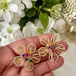 Puede incluir: Un par de pendientes en forma de flor hechos con alambre metálico dorado. Los pétalos están formados por finos hilos de alambre enrollado. El centro de cada diseño floral está adornado con un grupo de pequeñas cuentas redondas multicolores en tonos rojo, azul, verde y naranja.