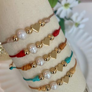 Puede incluir: Cinco pulseras de cordón ajustables en beige, rojo, mostaza, verde azulado y bronceado. Cada pulsera presenta un dije de corazón dorado central, dos pequeñas cuentas esféricas doradas y una perla de agua dulce blanca dispuestas simétricamente.