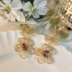 Puede incluir: Un par de pendientes de alambre en tono dorado con un diseño de doble flor. Cada pendiente consta de dos formas florales superpuestas hechas de alambre de oro fino, con el centro de la flor más grande adornado con pequeños cristales facetados.