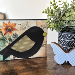 May include: A black bird with a yellow beak and a brown and white polka dot pattern on its wing is painted on a wooden sign. The sign has a floral design with a large orange flower and green leaves. The sign is sitting on a wooden table next to a blue and white gingham butterfly.