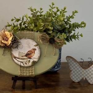 May include: A green oval wooden sign with a bird and floral design, a burlap bow, and a small easel. The sign is decorated with a brown and white checked butterfly.