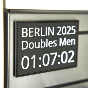Könnte beinhalten: Ein schwarzes rechteckiges Patch mit weißem Text "BERLIN 2025 Doubles Men 01:07:02". Das Patch ist an den Rändern genäht und auf einer Edelstahloberfläche montiert.