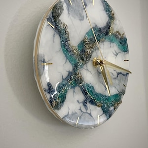 May include: A round wall clock featuring a white base with marbled gray veins and a textured, crystalline center section in shades of teal, deep blue, and gold. The clock has a thin gold-colored rim and slender gold-colored hands for the time.